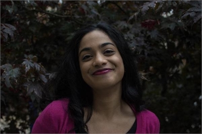 [ai] A woman smiling softly in front of a backdrop of dark leaves, wearing a pink cardigan over a black top.